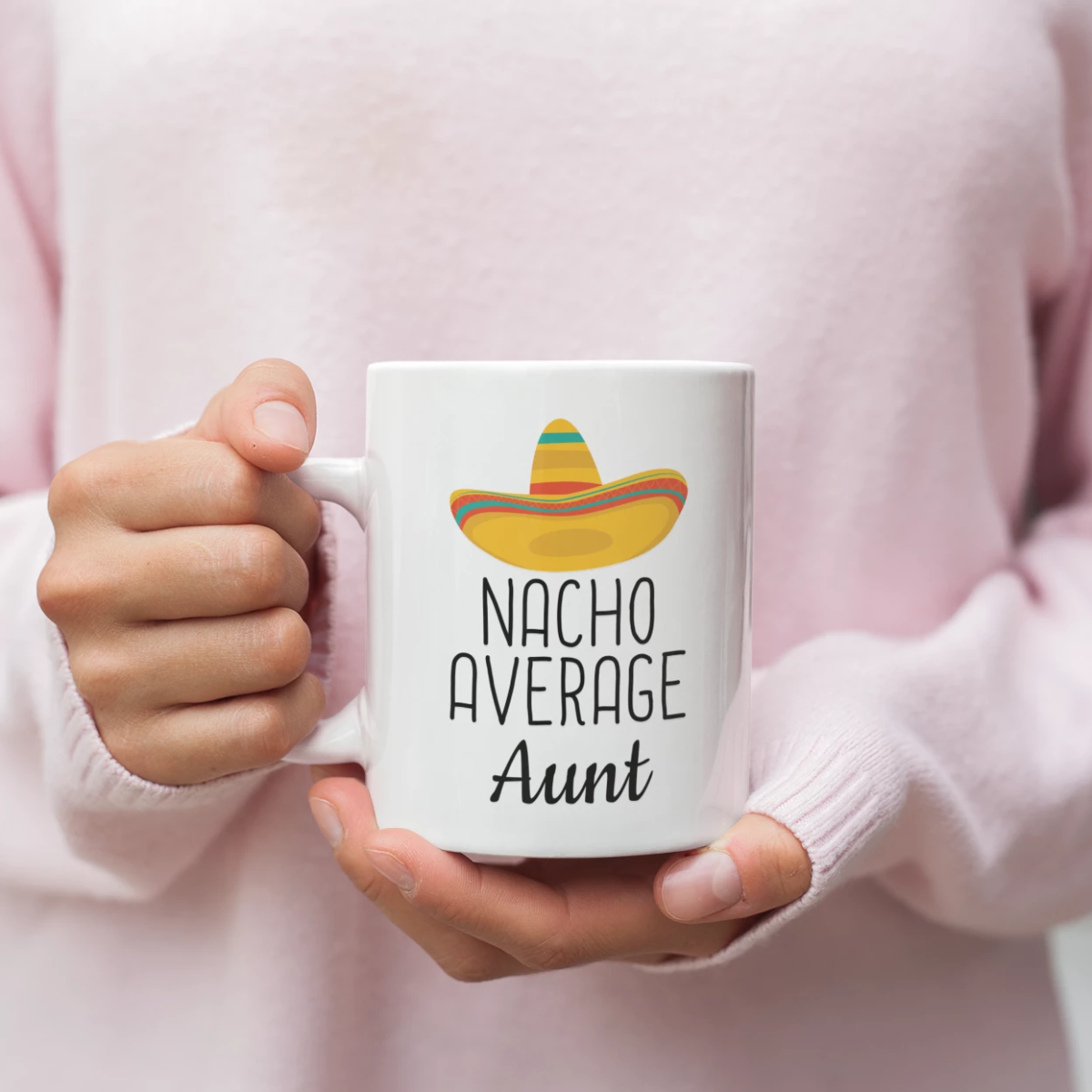 Person in a pink sweater holding a Nacho Average Aunt white mug