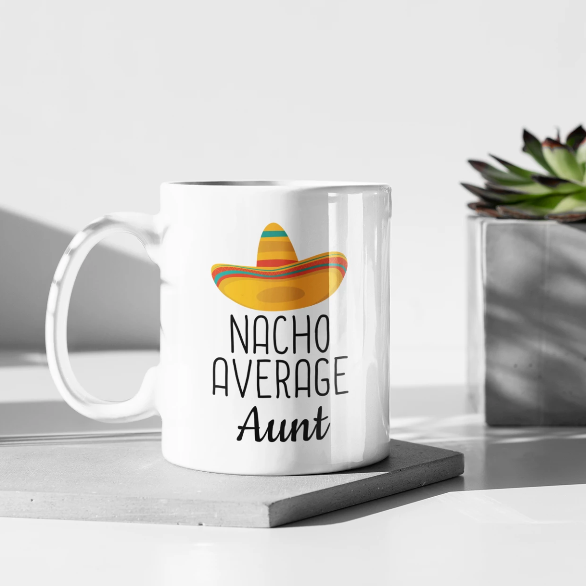 Nacho Average Aunt white mug on a square slab beside a potted succulent
